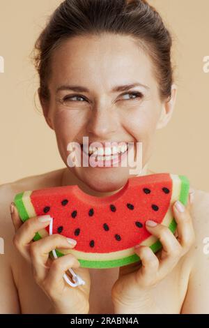 Glückliche 40-jährige Frau mit Wassermelonen-Duschschwamm auf beigefarbenem Hintergrund. Stockfoto