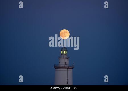 Der Buckmond, der am Himmel über einem Leuchtturm aufgeht, ist im Hafen von Malaga zu sehen. Anfang Juli findet der Vollmond der Hirsche als eines der beeindruckendsten astronomischen Ereignisse des Monats statt. Wenn der Vollmond stattfindet, erscheint er größer und heller als normal. Stockfoto