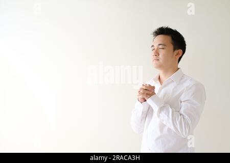 Ein junger asiatischer Mann, der ein weißes Hemd trägt und betet und sich nach rechts richtet. Isolierter weißer Hintergrund. Stockfoto