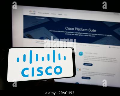 Person, die ein Mobiltelefon mit dem Logo der Aemrican Communications Company Cisco Systems Inc. Auf dem Bildschirm vor der Webseite hält. Konzentrieren Sie sich auf das Display des Telefons. Stockfoto
