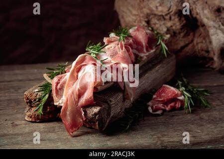 Italienischer Prosciutto Crudo oder spanischer Jamon mit Rosmarin auf altem Holzhintergrund. Stockfoto