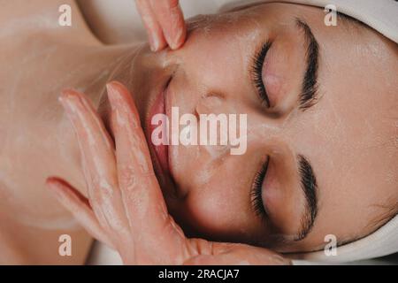 Crop anonyme Frau mit Gesichtsmaske auf Gesicht einer jungen Frau mit geschlossenen Augen in einem Spa-Salon. Stockfoto
