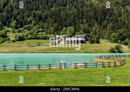 Holzzaun entlang des wunderschönen kleinen Bergsees von Brusson als ländliche Häuser und Bäume im Hintergrund im Aosta-Tal, Italien. Stockfoto