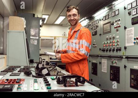 Junger Offshore-Offizier, der die Ausrüstung im Maschinenkontrollraum überprüft, mit lächelndem Gesicht. Stockfoto