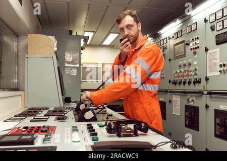 Offshore-Ingenieur kommuniziert über UKW-Funk mit Decksoffizier. Junger Marineexperte, der Funkprüfungen im Maschinenkontrollraum durchführt. Stockfoto