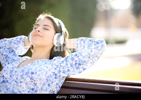 Eine glückliche Frau, die Musik hört, ruht sich auf einer Bank in einem Park aus Stockfoto