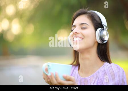 Glückliche Frau mit Kopfhörern, die Musik hört, die Kaffeetasse in einem Park hält und frische Luft atmet Stockfoto