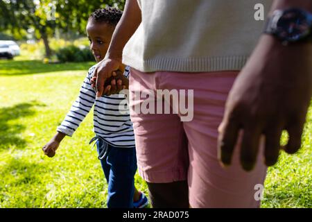 Der mittlere Teil des afroamerikanischen Vaters, der die Hand seines Sohnes hält und im Park spaziert Stockfoto