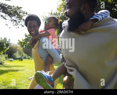 afroamerikanische fröhliche Eltern, die ihren Sohn und ihre Tochter auf dem Weg in den Garten mit dem Huckepack versorgen Stockfoto