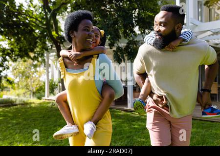afroamerikanische fröhliche Eltern, die Kinder beim Spazierengehen im Garten mit dem Huckepack betreten Stockfoto