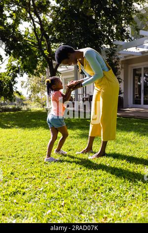 Seitenansicht einer afroamerikanischen Mutter, die ihre Tochter an den Händen hält, während sie auf einem grasbedeckten Feld im Hof steht Stockfoto