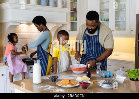 afroamerikanischer Vater und Sohn mischen Teig in die Schüssel, während Mutter und Tochter Pfannkuchen kochen Stockfoto