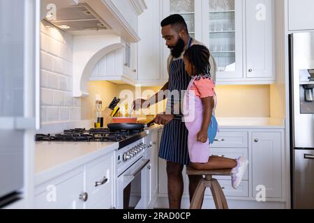 Eine afroamerikanische Tochter sieht Vater an, der Pfannkuchen auf dem Herd in der Küche kocht Stockfoto