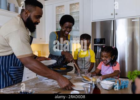 Fröhliche afroamerikanische Eltern servieren verspielten Kindern auf der dreckigen Küchenkinsel Pfannkuchen Stockfoto