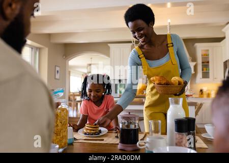 Lächelnde afroamerikanische Mutter, die der Tochter Pfannkuchen servierte und am Esstisch saß Stockfoto