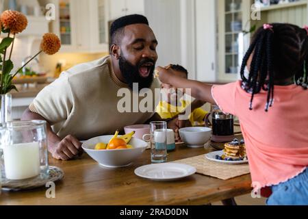 Ein afroamerikanisches Mädchen füttert den Vater mit Pfannkuchen, während es am Esstisch frühstückt Stockfoto