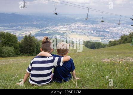 Zwei junge Freunde umarmen sich, während sie am Sommertag auf dem Berghang sitzen und die Landschaft genießen. Bruder, Liebes. Freundschaftskonzept. Rückansicht. Aktiv Stockfoto