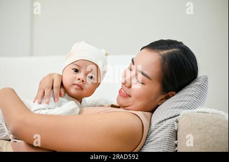 Wunderschöne asiatische Mutter hält ihren kleinen Jungen fest, während sie sich auf dem Sofa entspannt und Zeit mit ihrem Sohn verbringt. Stockfoto