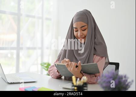Junge muslimische Asiatin liest Bücher, während sie in ihrem Büro arbeitet. Das Konzept der Geschäftsleute Stockfoto
