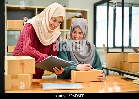 Eine glückliche junge asiatische muslimische Online-Verkäuferin, die mit ihrem Mitarbeiter im Büro arbeitet. KMU, Inhaber eines Online-Shops Stockfoto