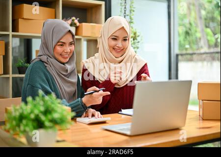 Eine glückliche junge asiatische muslimische Online-Verkäuferin, die mit ihrem Mitarbeiter im Büro arbeitet. KMU, Inhaber eines Online-Shops Stockfoto