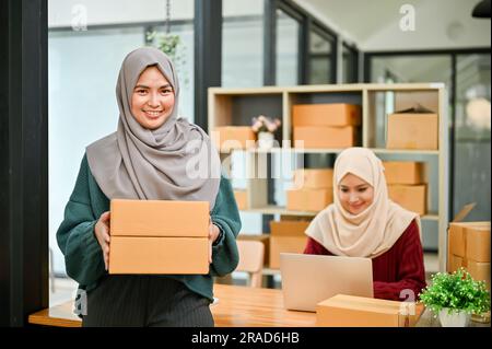 Eine glückliche junge asiatische muslimische Online-Verkäuferin, die mit ihrem Mitarbeiter im Büro arbeitet. KMU, Inhaber eines Online-Shops Stockfoto