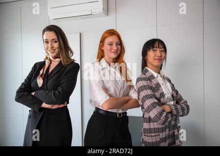 Drei glückliche Geschäftsfrauen mit zusammengefalteten Armen und selbstbewusster Ausdrucksform präsentieren sich im Büro Stockfoto