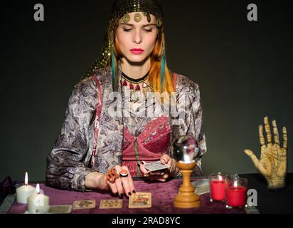 Erwachsenenfokussierte weibliche Wahrsagerin, die Tarotkarten liest, am Tisch mit Kerzen und magischer Kristallkugel Konzept der Enthaltsamkeit, Astrologie und Esote Stockfoto