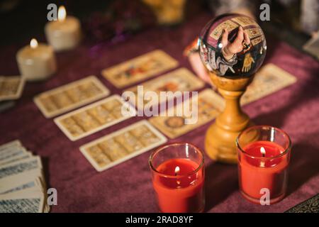 Selektiver Fokus der Kristallkugel, die das Erntegut reflektiert und die Zukunft vorhersagt, mit Tarotkarten, die sich in der Nähe brennender Kerzen auf dem Tisch vor unscharfem Hintergrund befinden Stockfoto