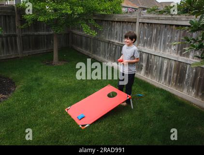 Ein glücklicher Junge, der ein Corn-Hole-Spiel in einem eingezäunten Garten spielt. Stockfoto