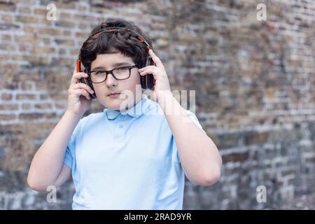 Porträt eines 11-jährigen Jungen mit Brille, kabelloser Kopfhörer, vor einer Steinwand, Musik hören, horizontal, Kopierraum Stockfoto