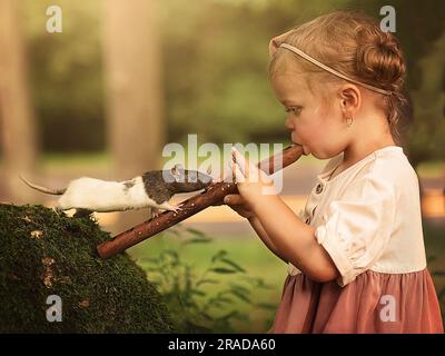 Ein kleines Mädchen, das draußen Rekorder spielt, während eine Ratte oder Maus sie beobachtet Stockfoto