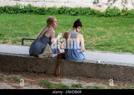 Zwei junge Frauen sitzen zusammen an einer Wand in einem Park in Split, kroatien. Freunde sitzen auf einer niedrigen Mauer zusammen, junge Frauen sitzen auf einer Mauer und entspannen sich. Stockfoto