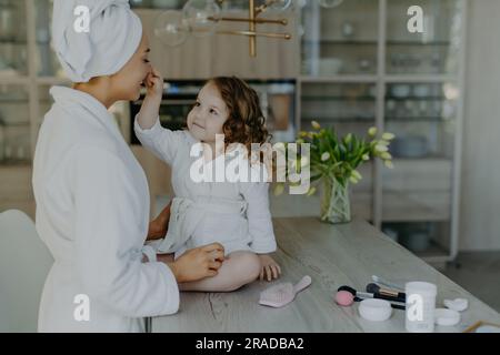 Das lockige Mädchen berührt mamas Nase, in weißem Bademantel, auf einem kosmetisch gefüllten Tisch, bereitet sich auf Make-up vor. Gemütliche Inneneinrichtung. Stockfoto