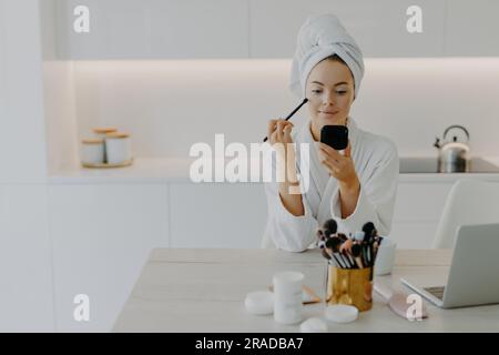 Erfrischte Frau trägt Gesichtspuder auf, benutzt Pinsel, posiert zu Hause in der Küche. Schreibtisch mit Kosmetikartikeln, Handtuch, Bademantel. Stockfoto