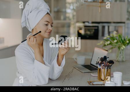 Schöne Frau trägt Make-up mit Pinsel auf, posiert zu Hause, vor dem Spiegel. Schreibtisch mit Laptop, Kosmetikgeräte, Bademantel. Selbstpflegekonzept. Stockfoto