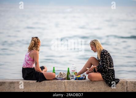 Zwei attraktive junge Frauen, die an der Wand am Meer sitzen und ein Picknick und ein paar Biere teilen, zwei Freunde, die an einer Hafenmauer sitzen und ein Bier und Essen teilen. Stockfoto