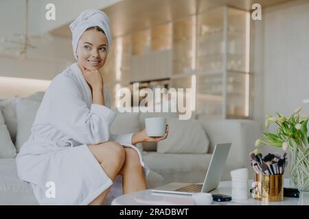 Fröhliche Frau im Bademantel, frische Haut, trinkt Kaffee, Puder, sitzt auf dem Sofa, Funktioniert auf Laptop, verwendet Kosmetik. Stockfoto