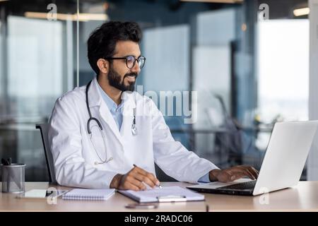 Erfolgreicher lächelnder Arzt arbeitet in einer modernen Klinikpraxis, hindu-Mann in weißer medizinischer Kittel arbeitet in einer Arztpraxis sitzt am Tisch mit Laptop. Stockfoto