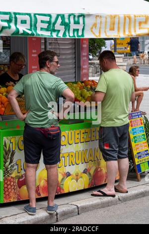 Zwei Männer stehen an einer Frischsafttheke in trogir, Croatiam-Männer kaufen Getränke an einem Marktstand und verkaufen frisch gepressten Orangensaft. Stockfoto