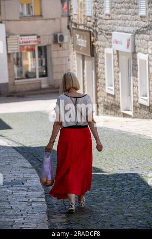 Eine elegante Dame, die auf der mediterranen Straße einkaufen geht, eine Dame mit rotem Rock, die einen Hügel hinuntergeht und eine Tasche trägt, eine stilvolle Dame mittleren Alters Stockfoto