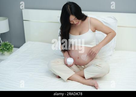 Eine schwangere Frau, die einen Streichelanfall hat, trägt herzförmige Creme auf ihren Bauch auf einem Bett auf Stockfoto