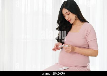 Junge Schwangere, die ein Blutzuckermessgerät in der Hand hält und zu Hause den Blutzuckerspiegel kontrolliert. Gestationsdiabetes-Konzept. Stockfoto