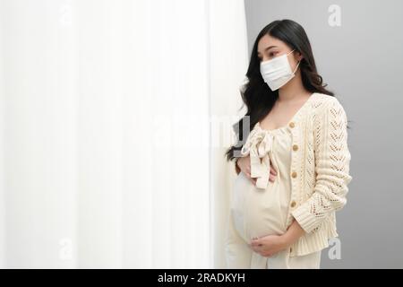 Schwangere Frau in medizinischer Maske und Blick durch Fenster in einem Schlafzimmer, bleiben Sie allein für Schutz Coronavirus (covid-19) Pandemie Stockfoto