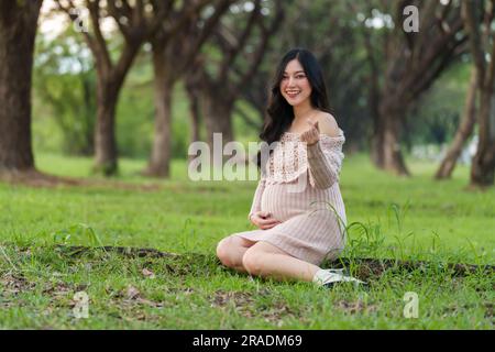 Glückliche schwangere Frau, die im Park sitzt und ihre Hände als Mini Hearts Symbol macht Stockfoto