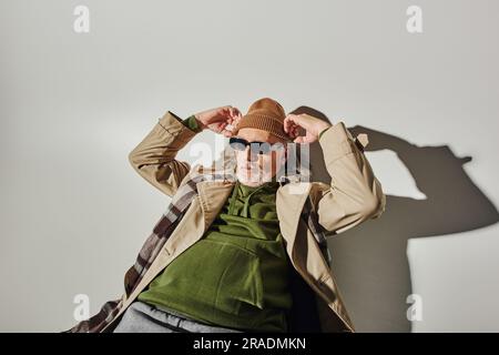 Ein alter Mann mit dunkler Sonnenbrille, grünem Hoodie und beigefarbenem Trenchcoat mit verstellbarer Mütze und Blick auf grauen Hintergrund mit Schatten, modisch und Stockfoto