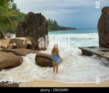 Wunderschöne Felsbrocken und weißer Sandstrand von intendance, eine Frau mittleren Alters, die zwischen Felsen und Wellen steht, Mahe Seychellen Stockfoto