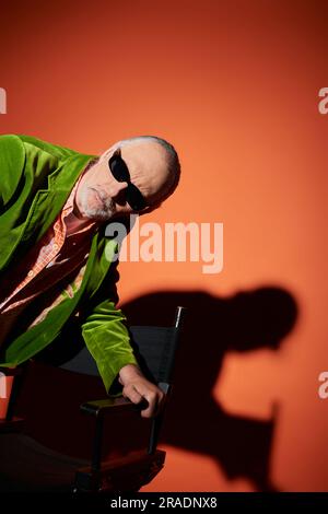 Ein rücksichtsvoller und neugieriger alter Mann in dunkler Sonnenbrille und grünem Velours-Blazer, der neben dem Stuhl steht und auf rot-orangefarbenen Hintergrund mit Schatten blickt Stockfoto