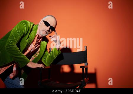 Ein rücksichtsvoller alter Mann mit dunkler Sonnenbrille und grünem Velours-Blazer posiert mit Stuhl und schaut auf die Kamera auf rotem und orangefarbenem Hintergrund mit Schatten, fashi Stockfoto