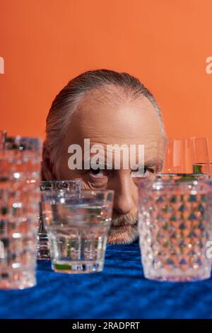 Porträt eines älteren grauhaarigen Mannes mit ausdrucksstarkem Blick auf die Kamera in der Nähe von Kristallgläsern voller klarem Wasser auf orangefarbenem Hintergrund, alternder Populat Stockfoto